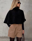 Woman wearing a black cape, beige shorts, and sunglasses against a gray background