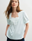 Woman wearing a light blue blouse with white lace details on a plain background