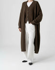 Person wearing a long brown cardigan over a white shirt and pants on a brown background