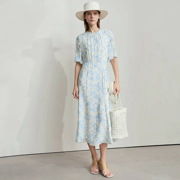 Woman wearing a light blue floral dress with a white hat and sandals, standing against a plain background.