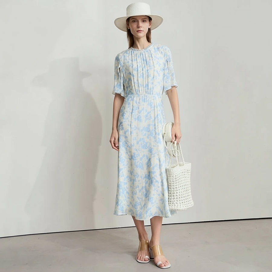 Woman wearing a light blue floral dress with a white hat and sandals, standing against a plain background.