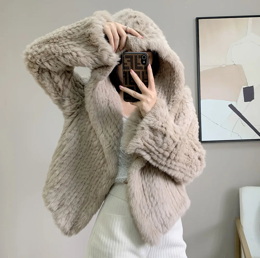 Person wearing a fluffy beige coat taking a mirror selfie in a room with a painting on the wall.