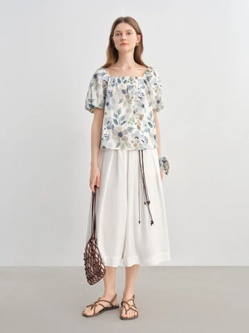 Woman wearing a floral top and white pants with a handbag on a plain background