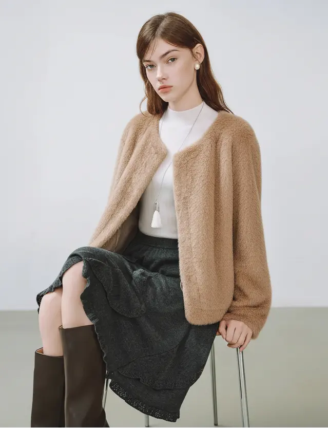 Woman wearing a beige cardigan over a white top and dark skirt, sitting on a chair with a plain background.