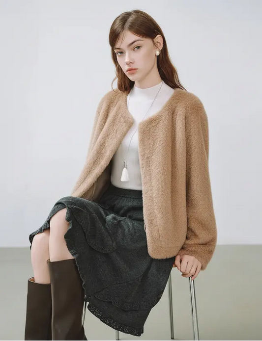 Woman wearing a beige cardigan over a white top and dark skirt, sitting on a chair with a plain background.