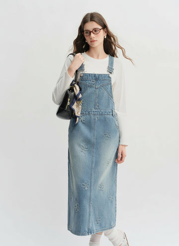 Woman wearing a denim pinafore dress with a white top and carrying a black bag on a light gray background.