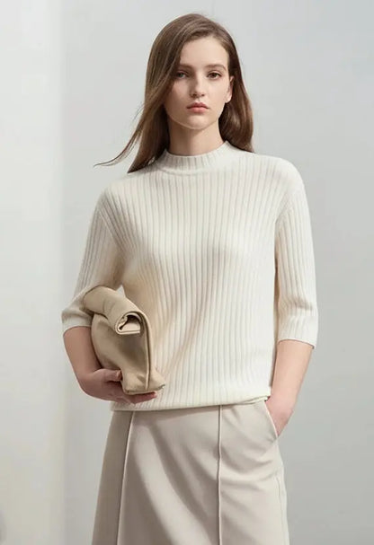 Woman wearing a white textured top and beige pants holding a beige clutch against a plain background
