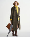 Person wearing a long brown coat with a yellow sweater, holding a brown bag on a white background
