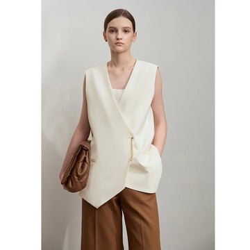 Woman wearing a white sleeveless top and brown pants holding a brown bag against a plain background