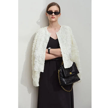 Woman wearing a white textured jacket over a black dress, holding a black handbag against a light gray background.