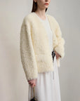 Woman wearing a light beige fluffy cardigan over a white dress against a plain background