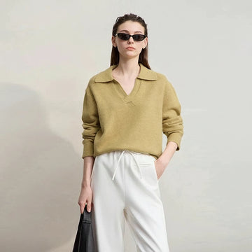 Woman wearing a green sweater and white pants with sunglasses, standing against a plain background. AMII brand visible.