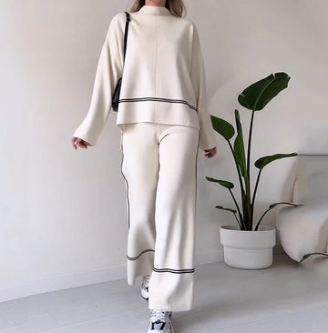 Person wearing a white outfit with black trim in a minimalistic room with a plant and sofa.