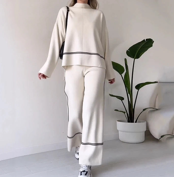 Person wearing a white outfit with black trim in a minimalistic room with a plant and sofa.