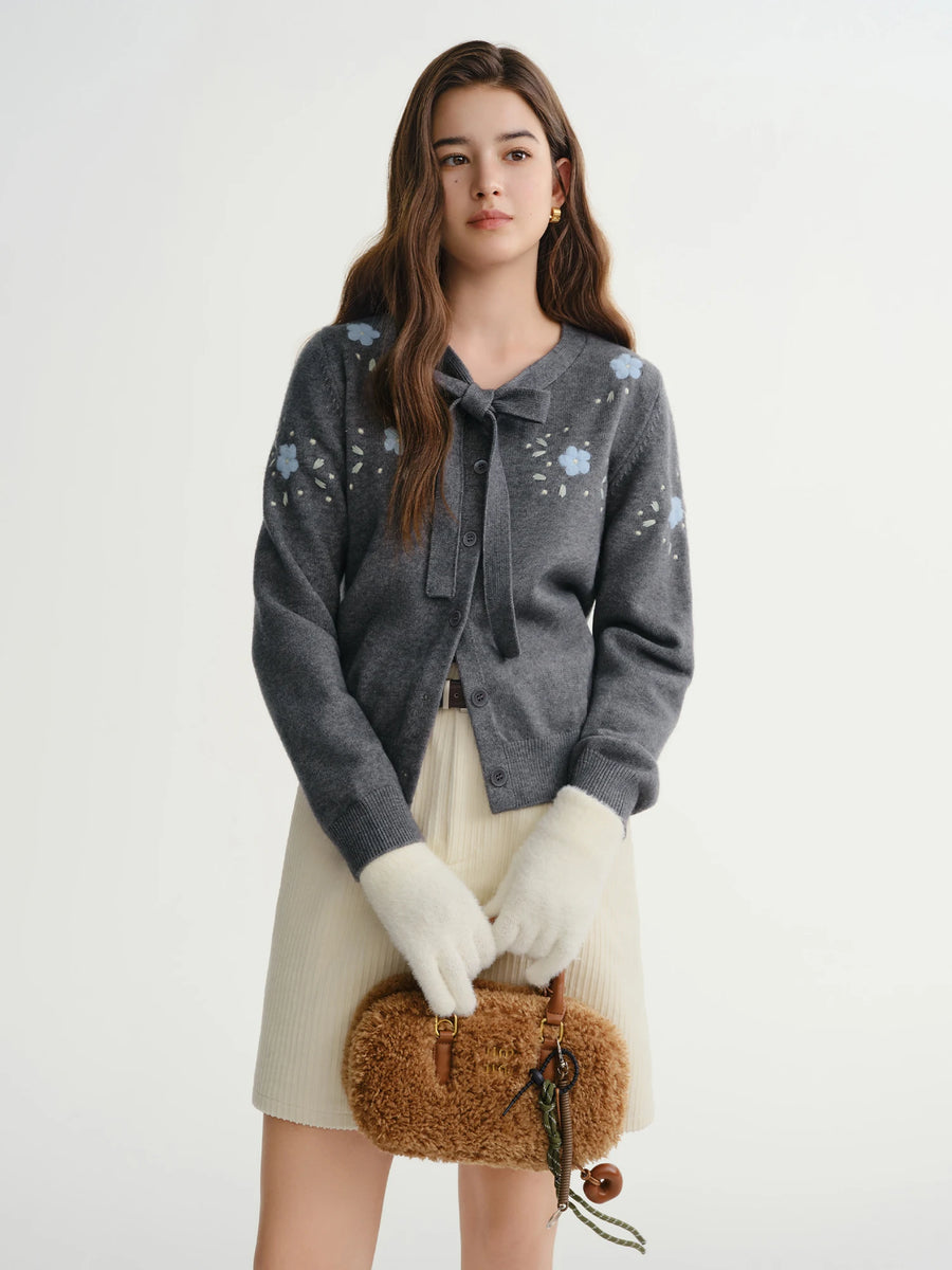 Woman wearing a gray floral cardigan, white gloves, and holding a brown bag on a white background