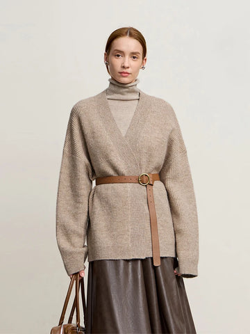 Woman wearing a beige sweater with a belt and holding a handbag on a plain background