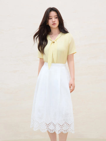 Woman wearing a yellow top and white skirt with lace details on a plain background