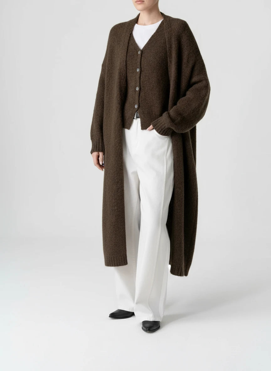 Person wearing a long brown cardigan over a white shirt and pants on a brown background