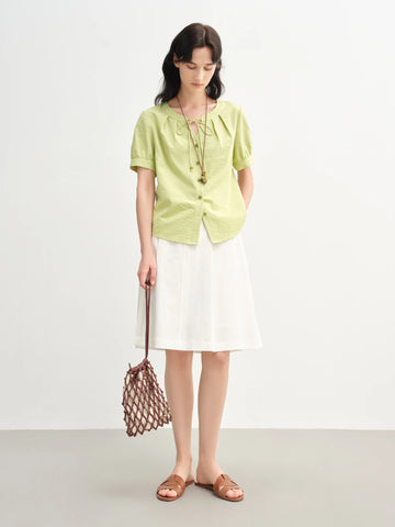 Woman wearing a light green shirt and white skirt holding a brown bag on a plain background