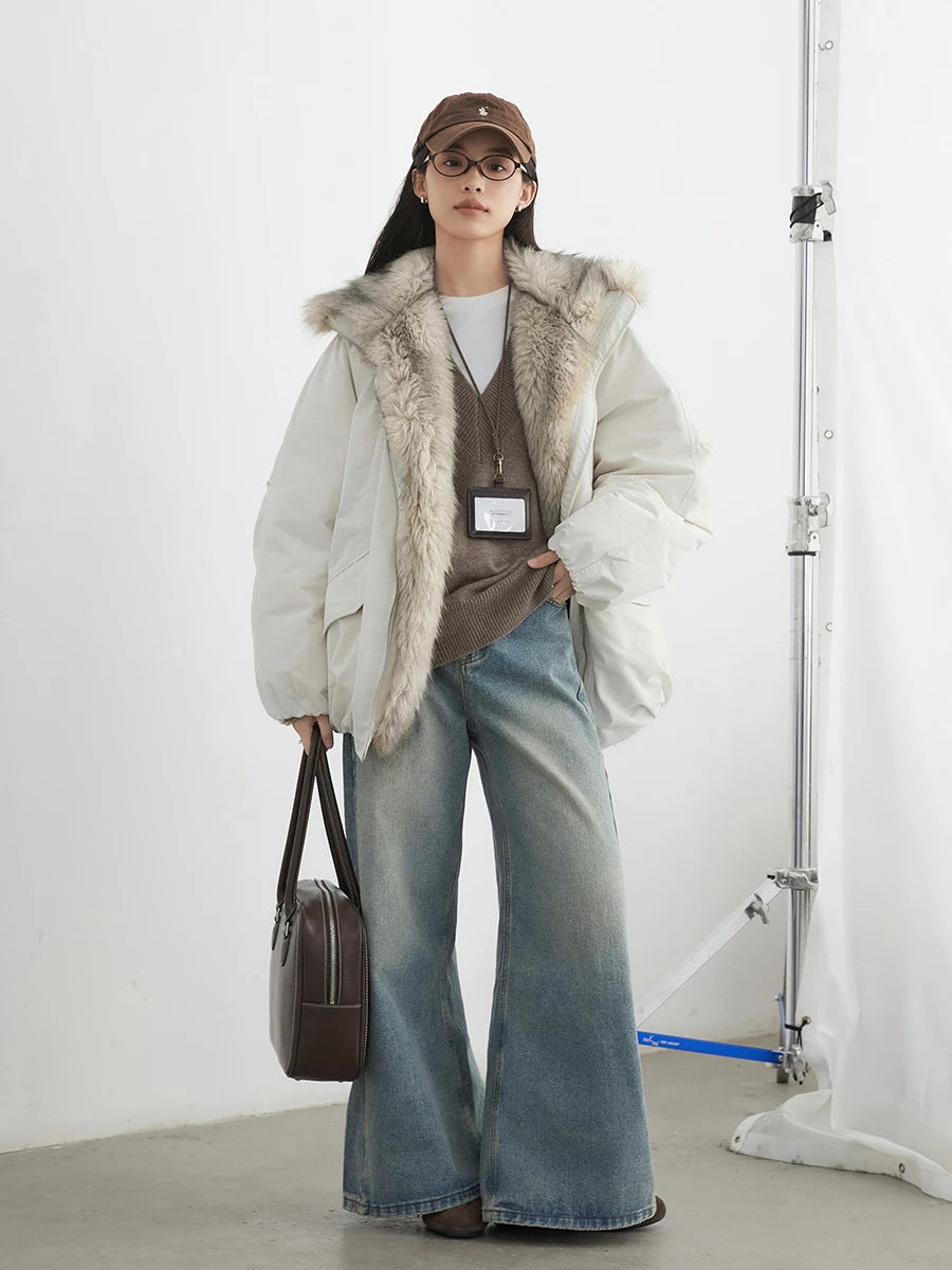 Person wearing a white puffer jacket with fur trim, brown sweater, blue jeans, and holding a brown bag against a white background.
