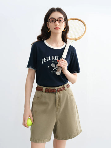 Woman holding a tennis racket and ball, wearing a navy blue t-shirt with text and beige shorts on a white background