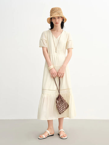 Woman wearing a light-colored dress and straw hat, holding a woven bag on a plain background