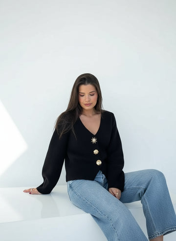 Woman wearing a black cardigan with gold buttons and light blue jeans standing outdoors.