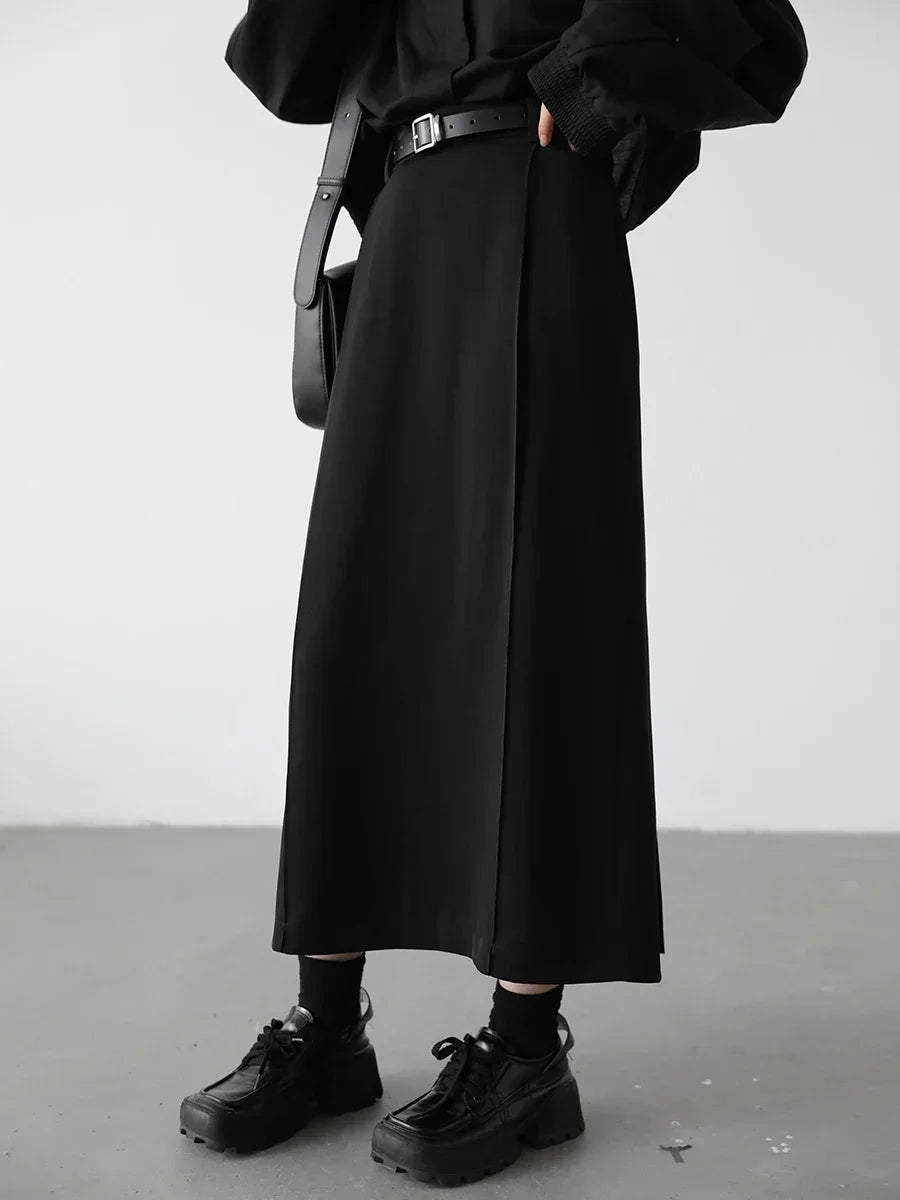 Person wearing a black outfit with a long black skirt, black shoes, and a black bag against a white background.