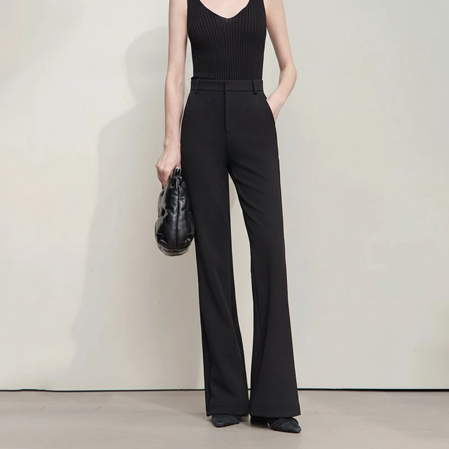 Woman wearing black high-waisted pants and a black top, holding a black handbag against a plain background.