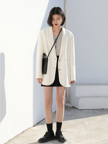 Woman wearing a white blazer and black skirt standing against a light-colored wall.