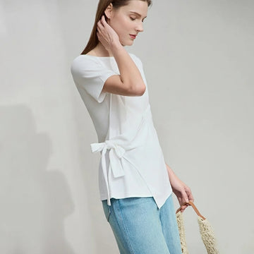 Woman wearing a white blouse with a bow detail and blue jeans, holding a straw bag on a plain background.