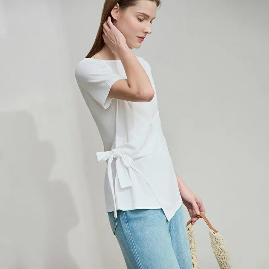 Woman wearing a white blouse with a bow detail and blue jeans, holding a straw bag on a plain background.