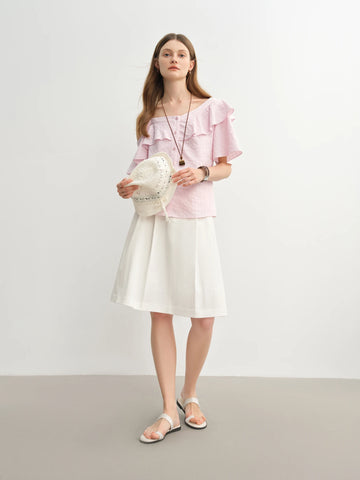 Woman wearing a pink top and white skirt holding a white bag against a plain background