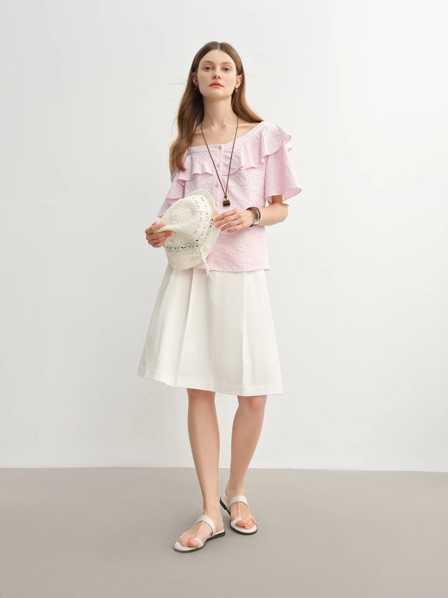 Woman wearing a pink top and white skirt holding a white bag against a plain background