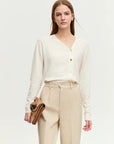 Woman wearing a white cardigan and beige pants holding a brown clutch against a white background