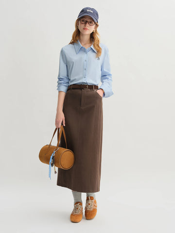 Woman wearing a light blue shirt, brown skirt, and holding a round brown bag on a white background