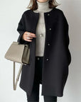 Person wearing a black coat holding a beige handbag on a plain background