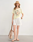 Woman wearing a yellow t-shirt with a peace symbol design, white shorts, and a straw hat, standing against a plain background.