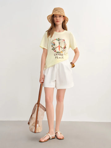 Woman wearing a yellow t-shirt with a peace symbol design, white shorts, and a straw hat, standing against a plain background.