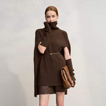 Woman wearing a brown ribbed sweater dress with a belt and holding a brown leather bag on a light gray background