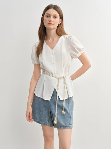 Woman wearing a white blouse with a tie waist and denim shorts on a plain background