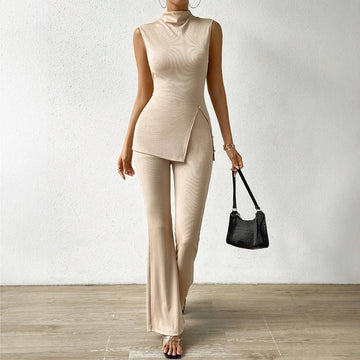 Person wearing a beige sleeveless top and pants holding a black handbag against a white wall.