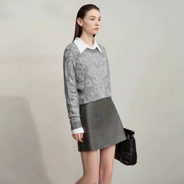 Woman wearing a gray sweater and black leather skirt with a white collar, holding a black bag on a plain background. Brand 'Amii' visible.