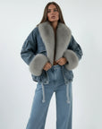 Person wearing a denim jacket with fur trim, sitting on a bed.