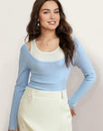 Woman wearing a light blue long-sleeve top over a white tank top with a beige skirt on a plain background